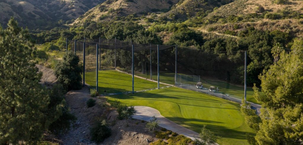 Golf driving range with tall netting, golf carts, and putting area surrounded by canyon hills at San Dimas Canyon Golf Course