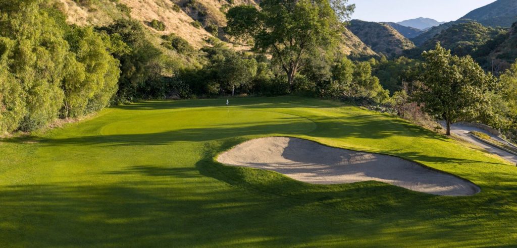 Golf green with white flag and curved sand bunker nestled in a tree-lined canyon at San Dimas Canyon Golf Course