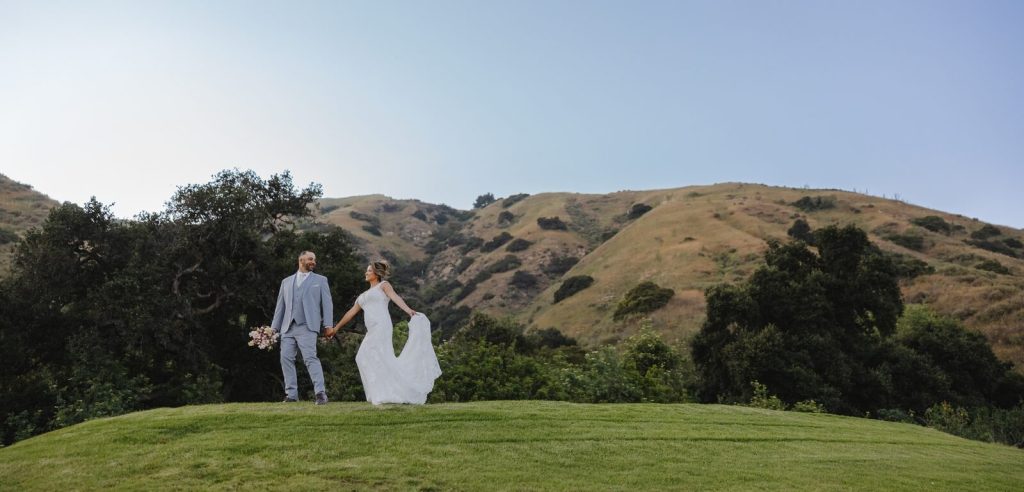 Bride and groom holding hands while walking across a grassy hilltop with mountain views at San Dimas Canyon Golf Course